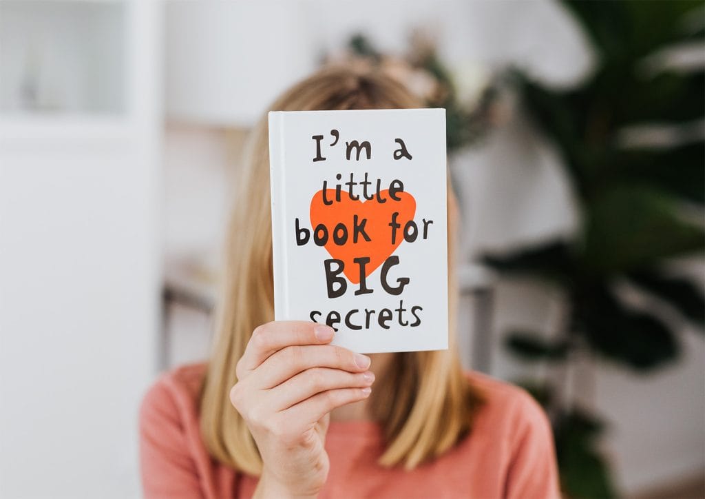 Image of a person holding up a small book covering their face. The typography used on the cover design is childlike and black. There is a red heart behind the text on a white background. Cover Design.
