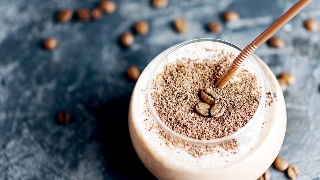 Close up image of a glass of espresso protein smoothing. There is a brown straw in the smoothie and coffee beans scattered on the table top.