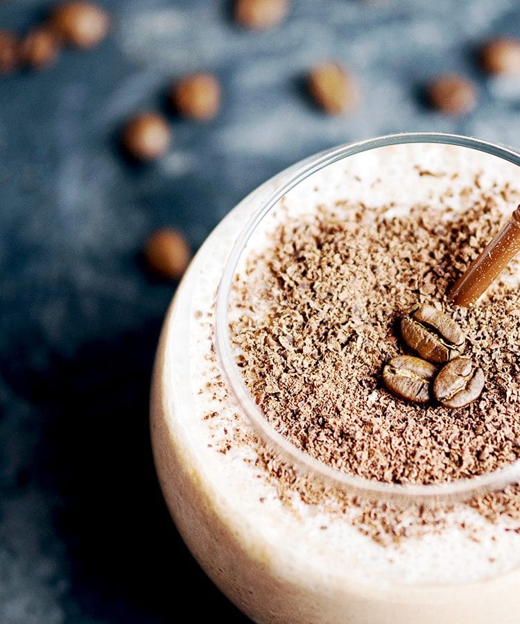 Close up image of a glass of espresso protein smoothing. There is a brown straw in the smoothie and coffee beans scattered on the table top.