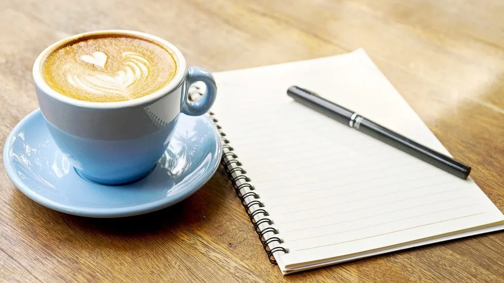 An image of a blue earthenware cup and saucer of coffee and an empty notebook and pen.