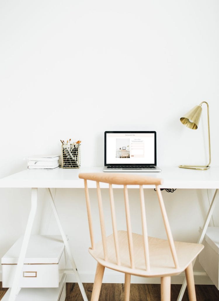Image of a white desk on top of which is a lamp, a laptop and a container of pens. There is also a beechwood ladder back chair.