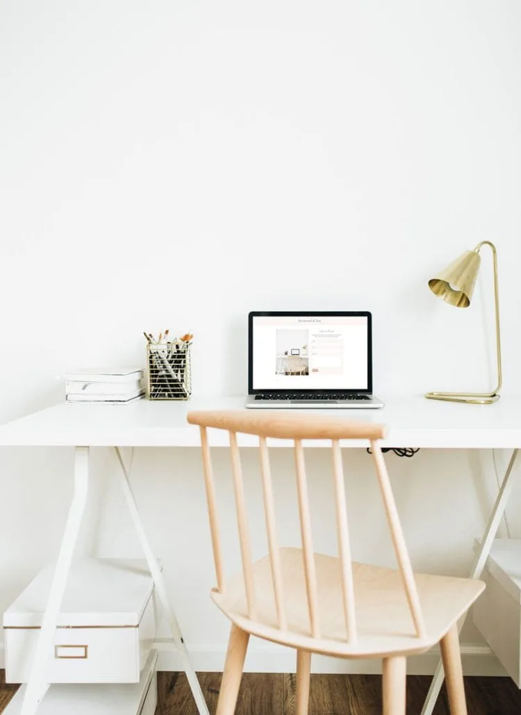 Image of a white desk on top of which is a lamp, a laptop and a container of pens. There is also a beechwood ladder back chair.