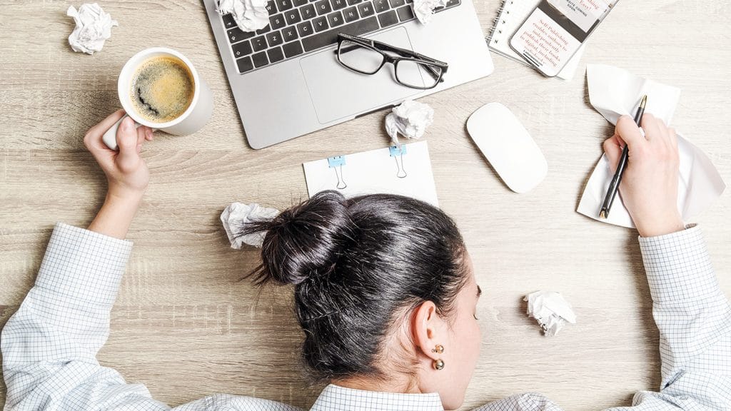 Image of a person asleep at their desk. There is a mug of what appears to be cold coffee, scrunched up paper strewn on desk, a laptop and mobile phone. Self-care.