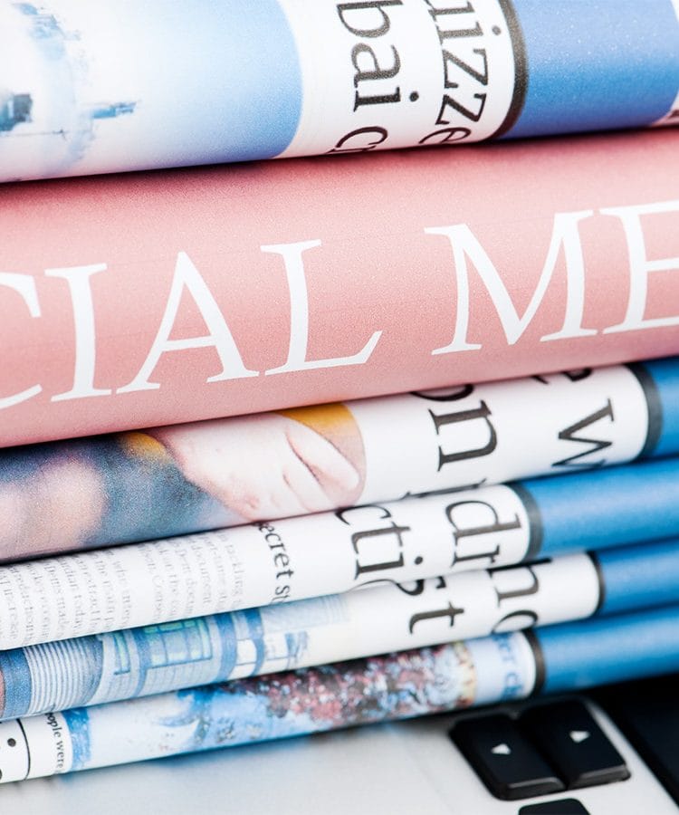 An image of a stack of folded newspapers on a laptop. In the middle of the stack, one can see a book's spine with the words Social Media.