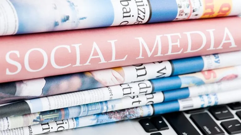 An image of a stack of folded newspapers on a laptop. In the middle of the stack, one can see a book's spine with the words Social Media.