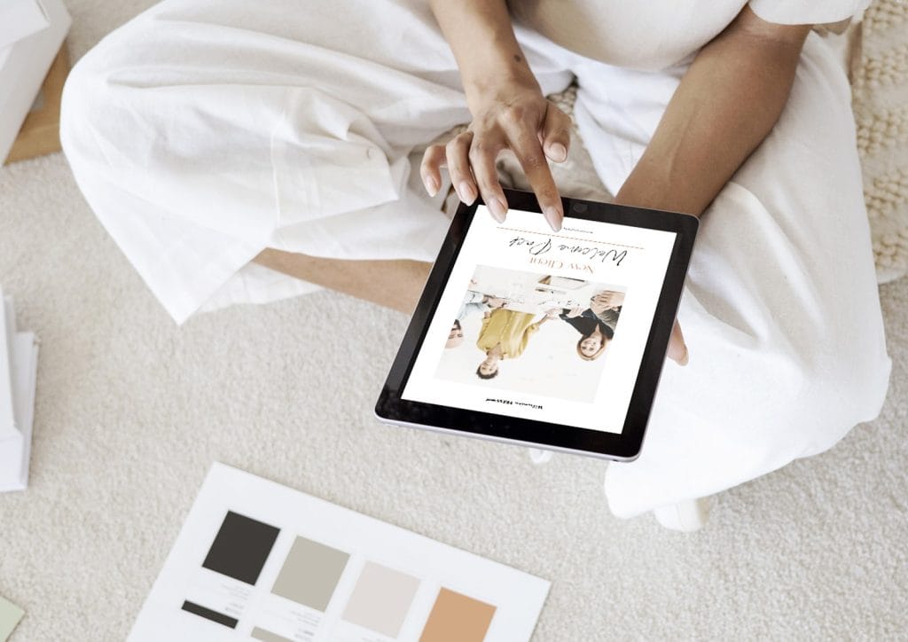 Image of a person sitting crossed legged on a white carpet holding a tablet and looking at the front cover of Wilsonian Press' New Client Welcome Pack.