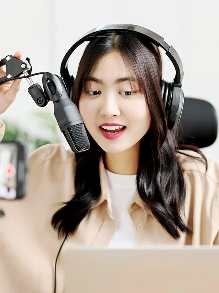 Image of a podcaster wearing headphones and holding an adjustable microphone, sits in front of a laptop and mobile phone camera producing a podcast.