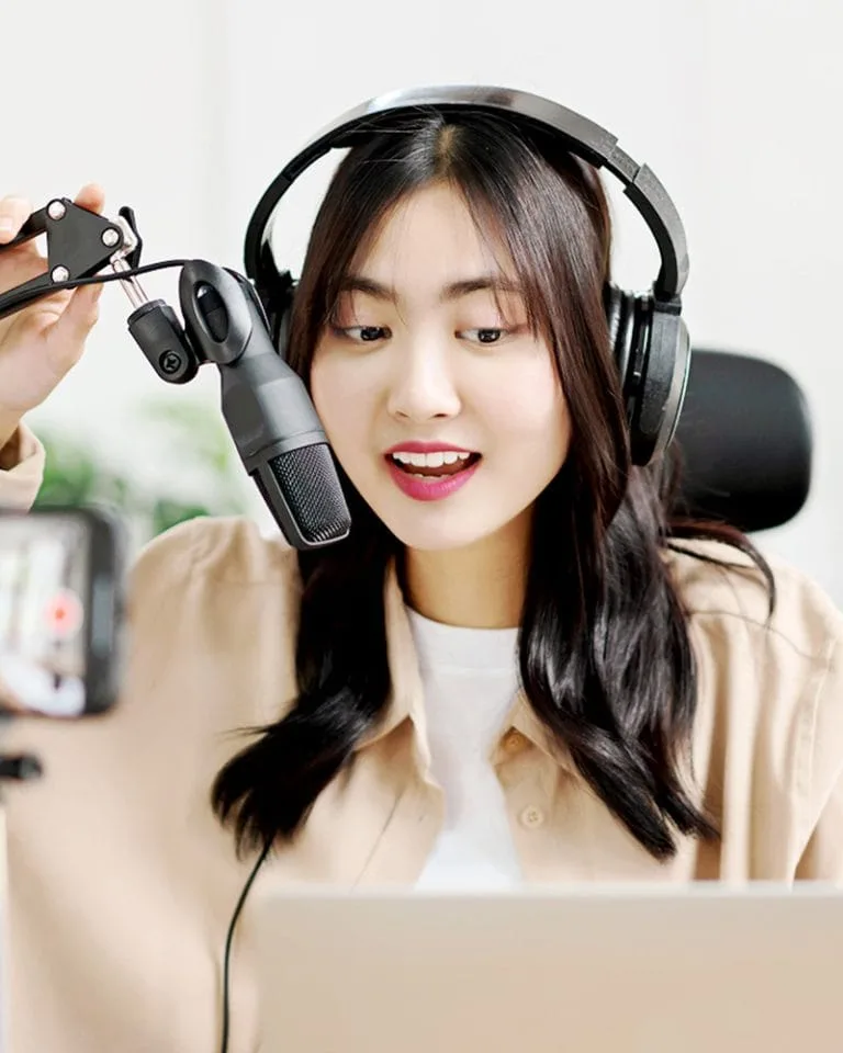 Image of a podcaster wearing headphones and holding an adjustable microphone, sits in front of a laptop and mobile phone camera producing a podcast.