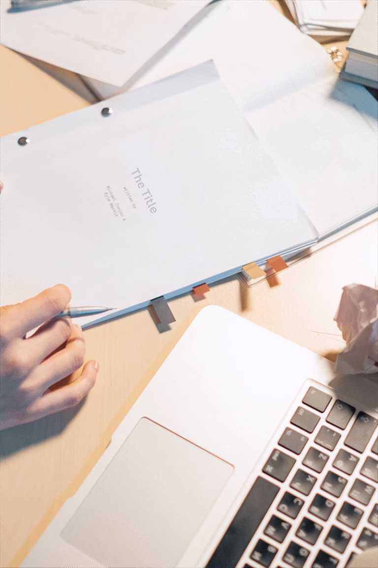 Image of a person editing a manuscript sat at a desk, the manuscript is seen being held. There is crumpled paper, a laptop and other clutter on the desk.